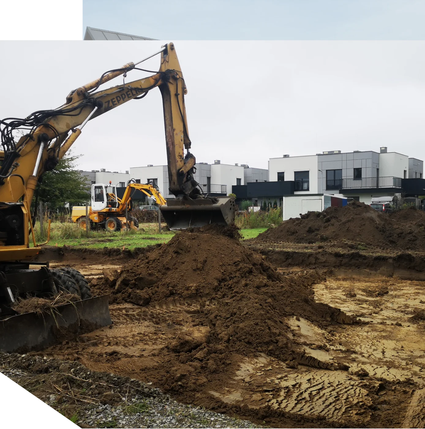 Koparka wykonująca wykop pod płytę fundamentową na terenie przyszłej inwestycji z widocznymi domami w tle