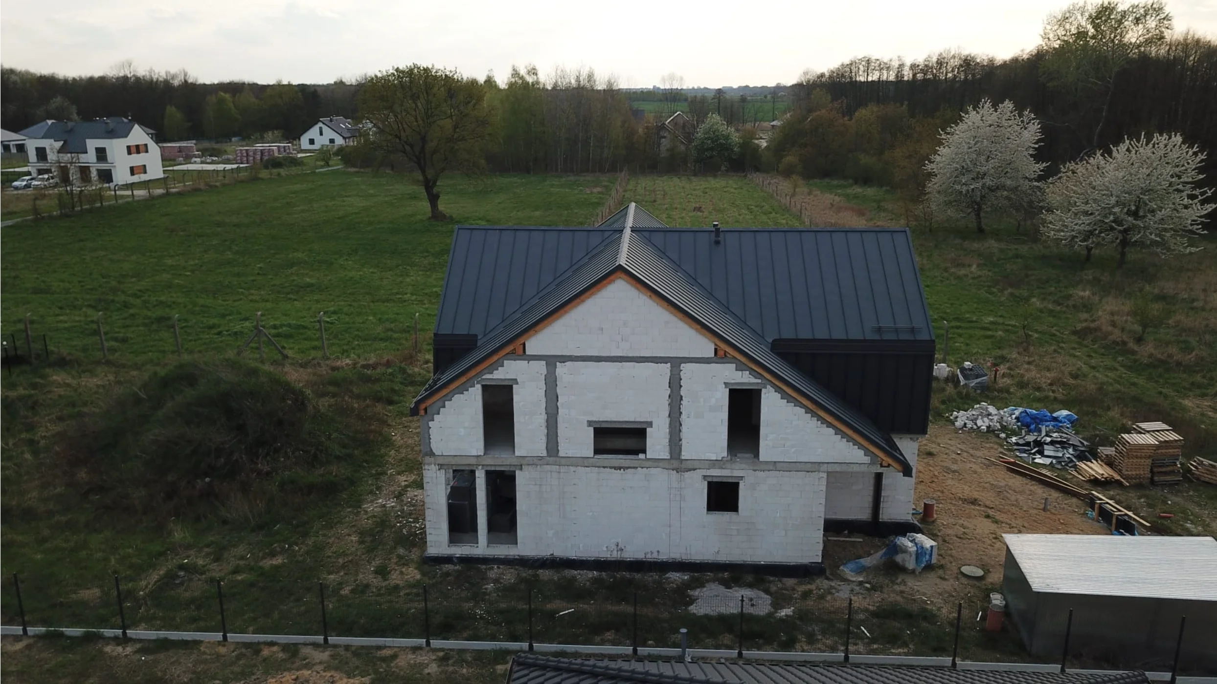 Dom w stanie surowym z zamontowanym dachem i widocznymi otworami okiennymi na tle zieleni