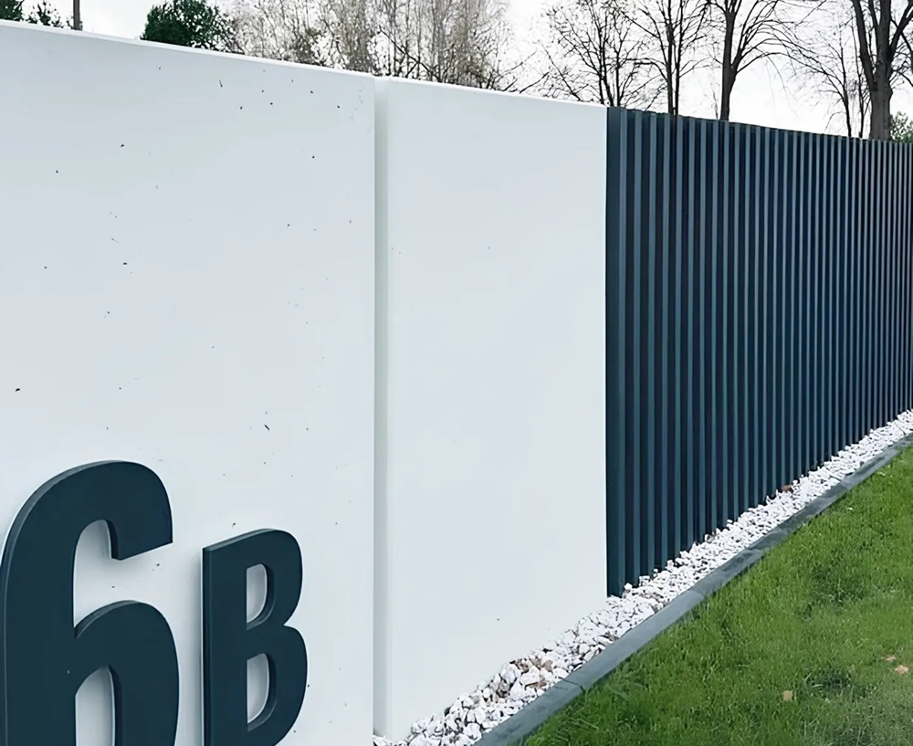 Białe ogrodzenie betonowe z ciemnymi pionowymi panelami oraz numerem domu, otoczonym otoczakami i trawnikiem